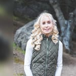 Senior woman with curly blonde hair smiling outdoors, wearing a white shirt and green vest, showcasing vitality and health